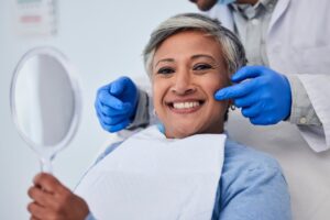 woman smiling, holding a mirror, and sitting in the dentist’s chair